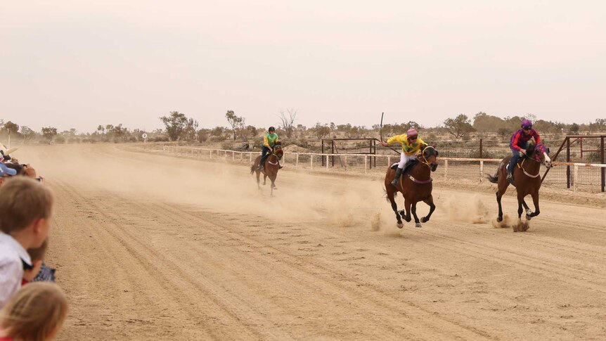 three horses with jockeys on them gallop on a dirt racecourse.  There are spectators.