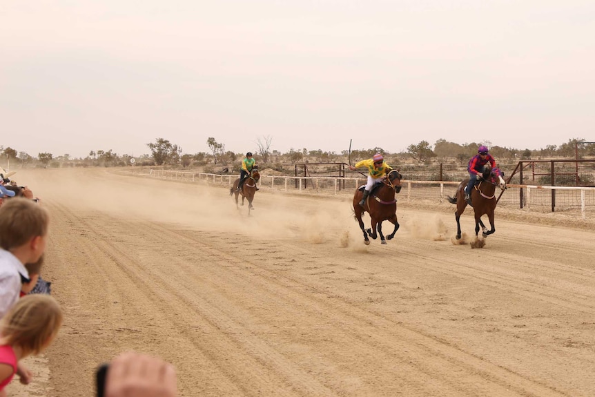 three horses with jockeys on them gallop on a dirt racecourse.  There are spectators.