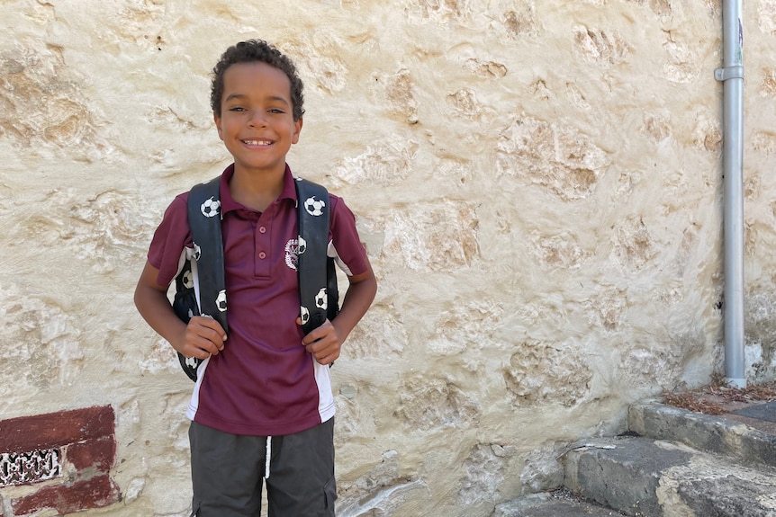 Tidiwa stands holding the straps of his school bag smiling in front of a brick wall