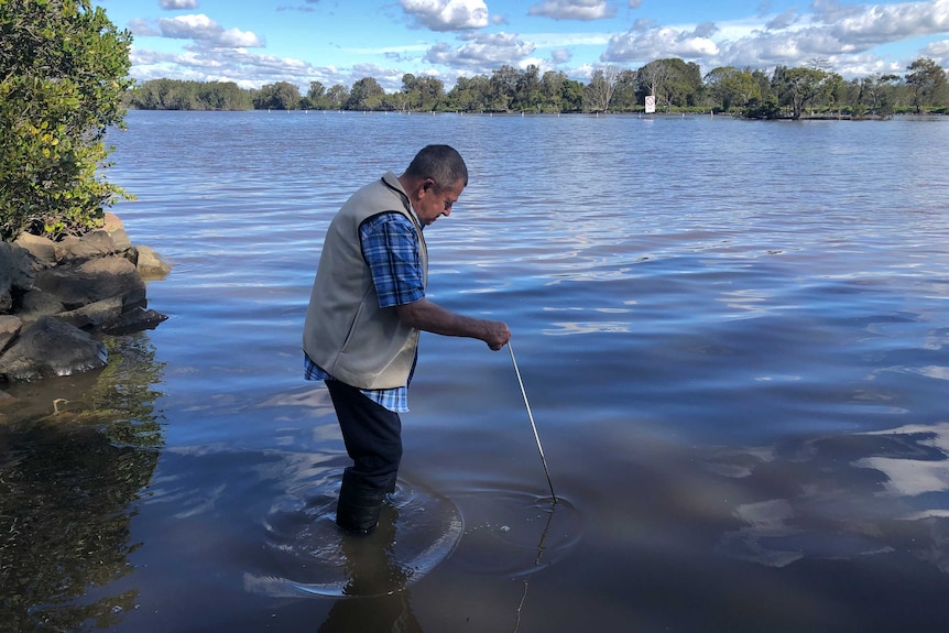 A man standing in shallow river water, holding a metal spike into the water.