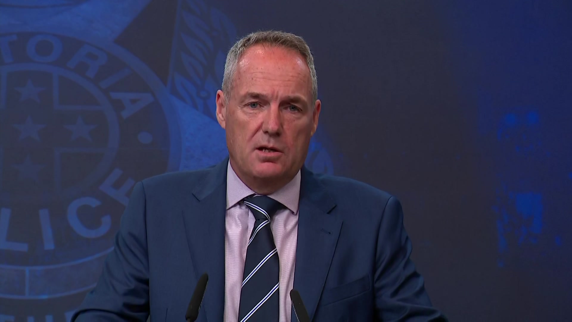 Un hombre de cabello gris con una chaqueta azul, una camisa de cuello blanca y una corbata a rayas azules y blancas se encuentra frente a una pantalla azul marino.
