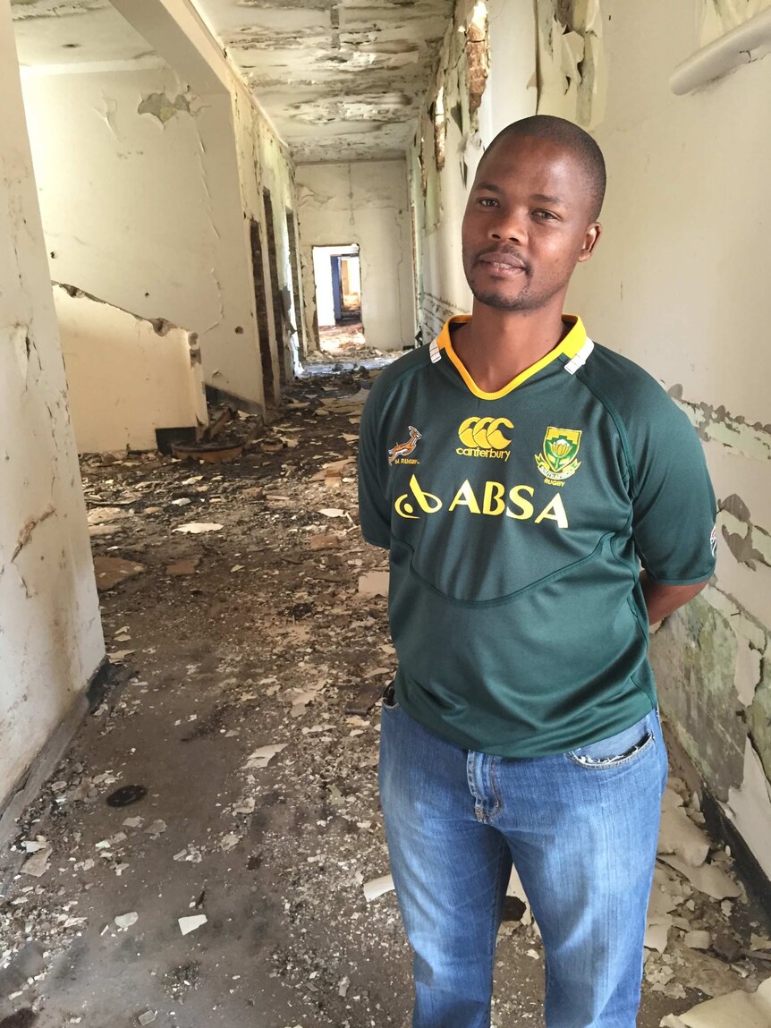Pule stands in a dilapidated building with paint peeling on the walls and debris strewn on the floor.