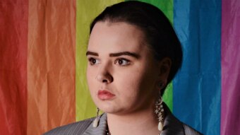 Kirra Hampson wears a grey pinstripe suit, earrings and a necklace and stands against a rainbow flag.