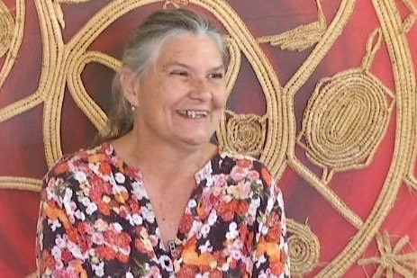 A woman is looking away from the camera smiling. There is a woven art piece with a red background behind her.