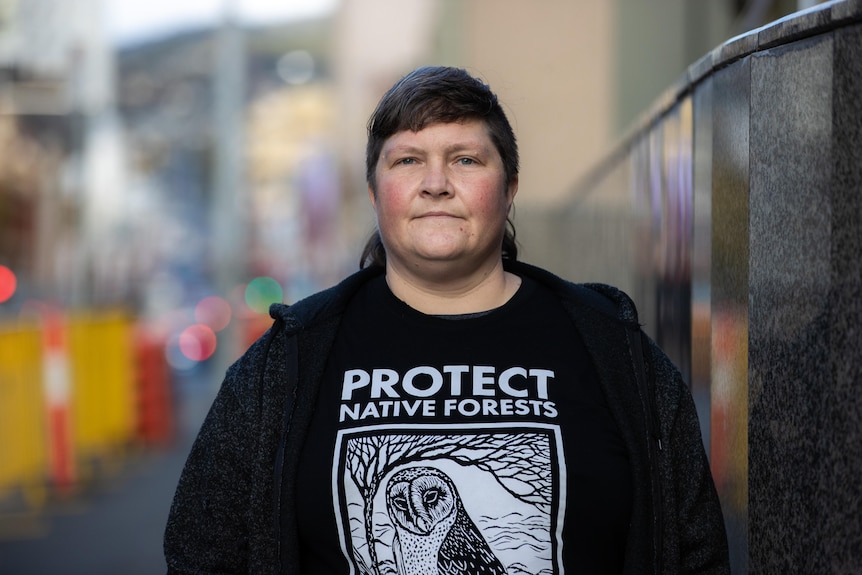 A woman with dark mullet haircut and black t-shirt with white owl picture and protect native forests slogan.