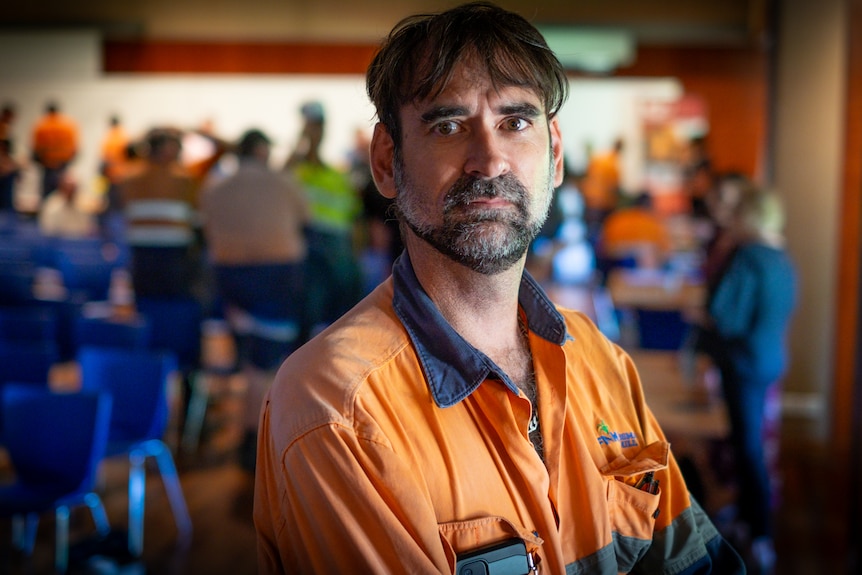 A dark-haired man in high-vis stands in a community hall, looking concerned.