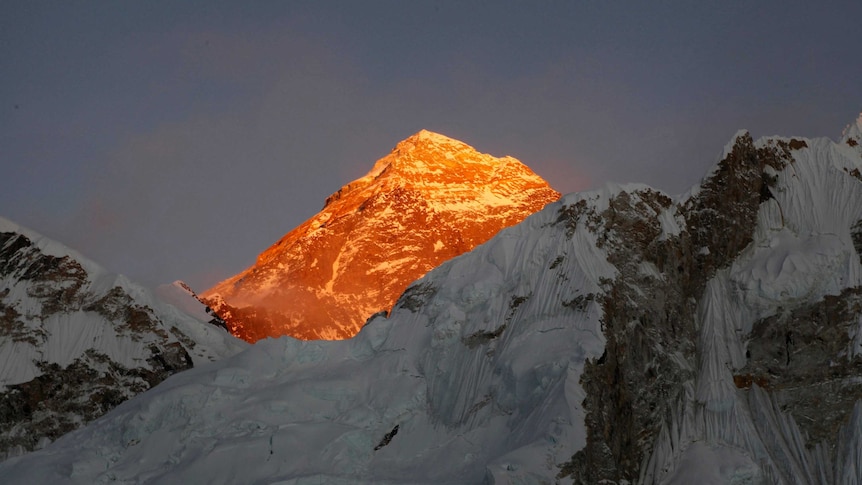 Mt. Everest glows brilliant orange in the sunlight behind a cold, blue peak.