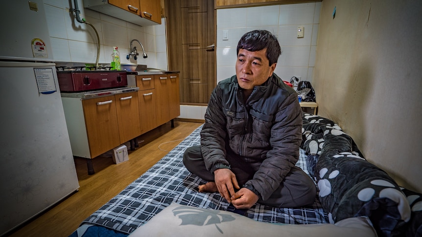 A middle-aged Asian  man sits on a bed next to a kitchenette looking forlorn.