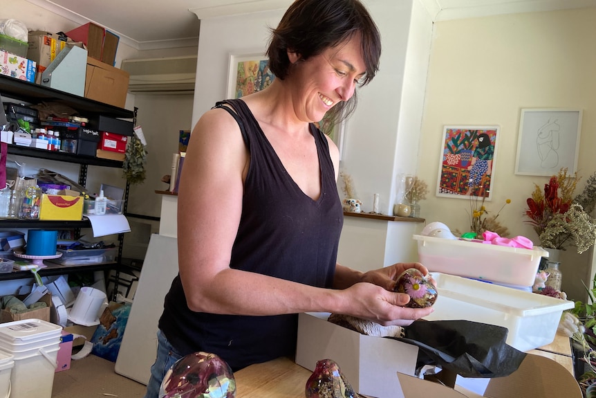 Woman holding a round resin art ornament in a home studio with a packing box on a bench in front of her.