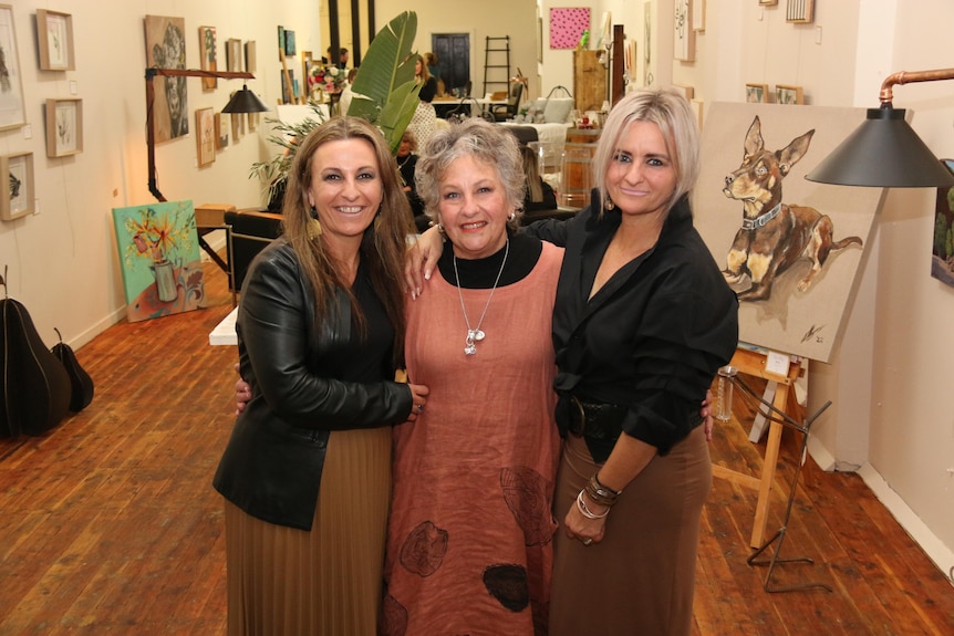 Three women stand smiling in an art gallery.