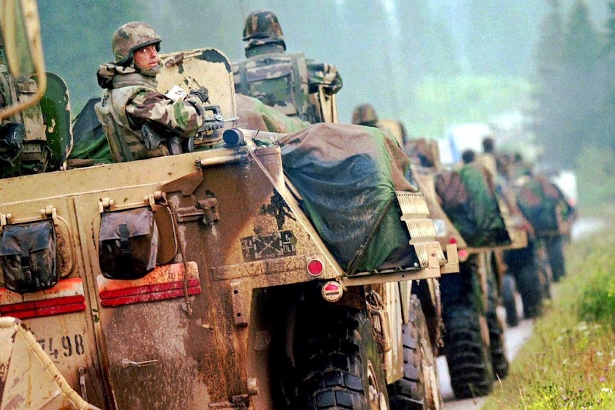 A convoy of armoured army vehicles drives down a road, with one soldier looking at the camera.