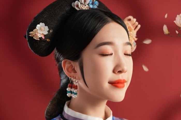 A woman standing in front of a red background dressed in traditional Chinese clothes closes her eyes.