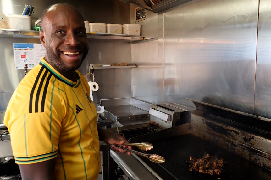 A Jamaican man cooks chicken on a grill inside a food truck.
