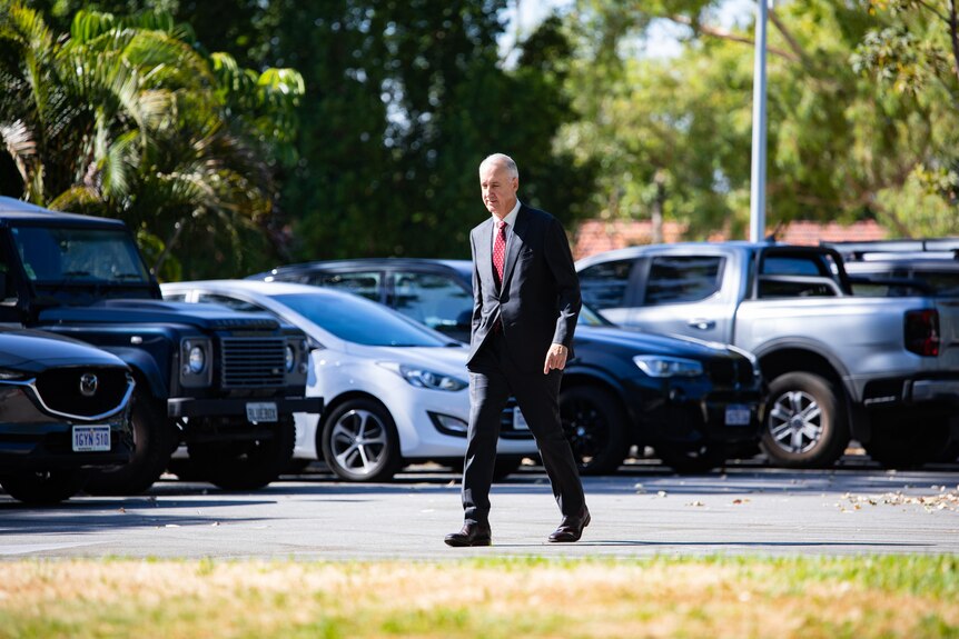 A man wearing a black suit and red tie walks through a carpark