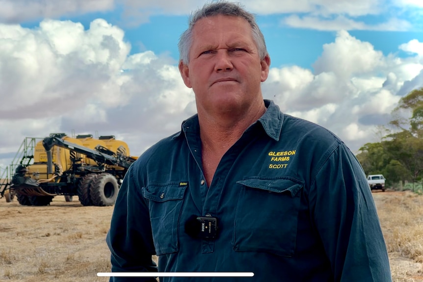 A grey-haired man wearing a long-sleeved, blue work shirt, stands in a grain field, with a tractor in the background.