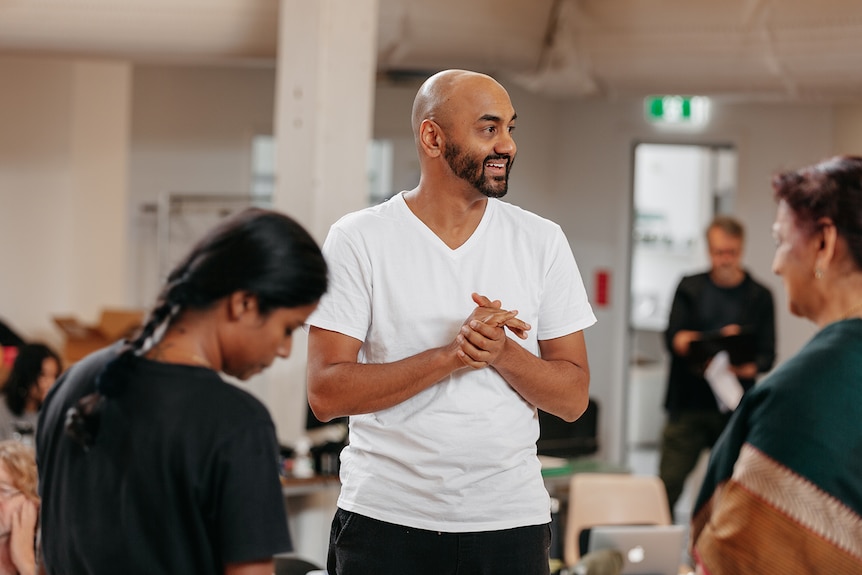 A Sri Lankan man in his early 40s, bald head, dark beard and white tshirt, stands with hands clasped, others in the foreground