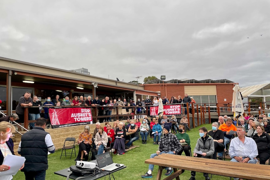 A crowd of people protesting powerlines stand or sit as they listen to a speech.