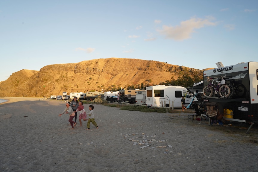 Four kids run across the sand smiling, and a row of parked caravans is parked behind them. Mountains are in the distance.