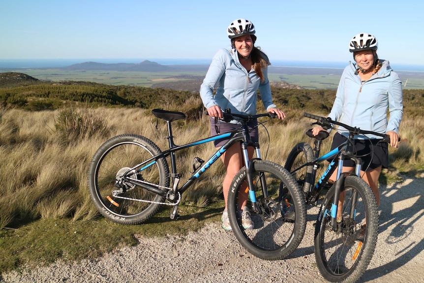 two women stand on top of a mountain range with their mountain bikes