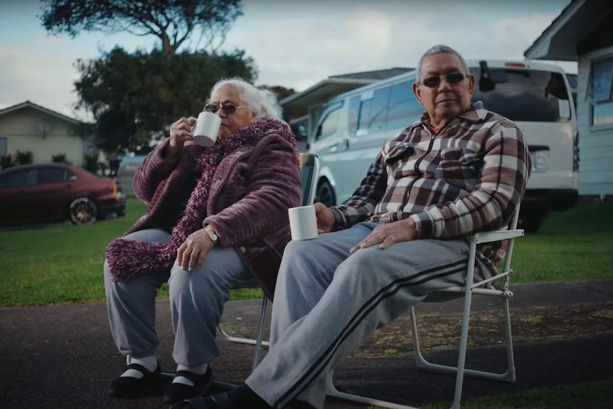 An elderly couple, Moeva and Ofisa Iaopo, sit in folding chairs on a suburban sidewalk with mugs in their hands.
