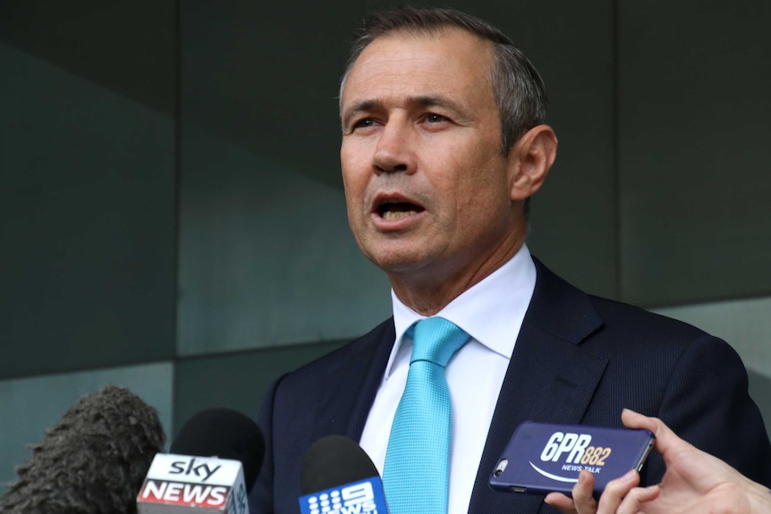 A picture of Roger Cook in blue tie speaking into microphones outside the ABC Perth office.