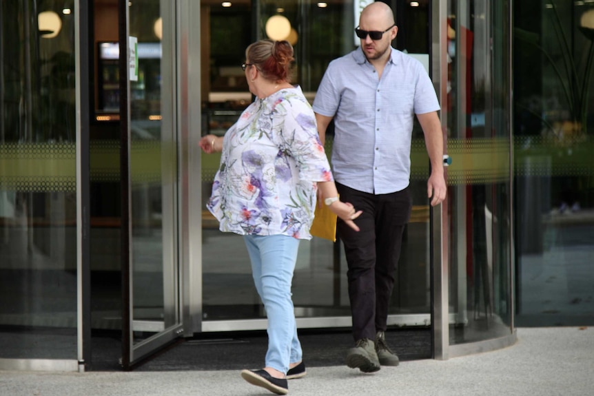 James walks out of the revolving door wearing sunglasses and holding an envelope, a woman walking ahead of him.