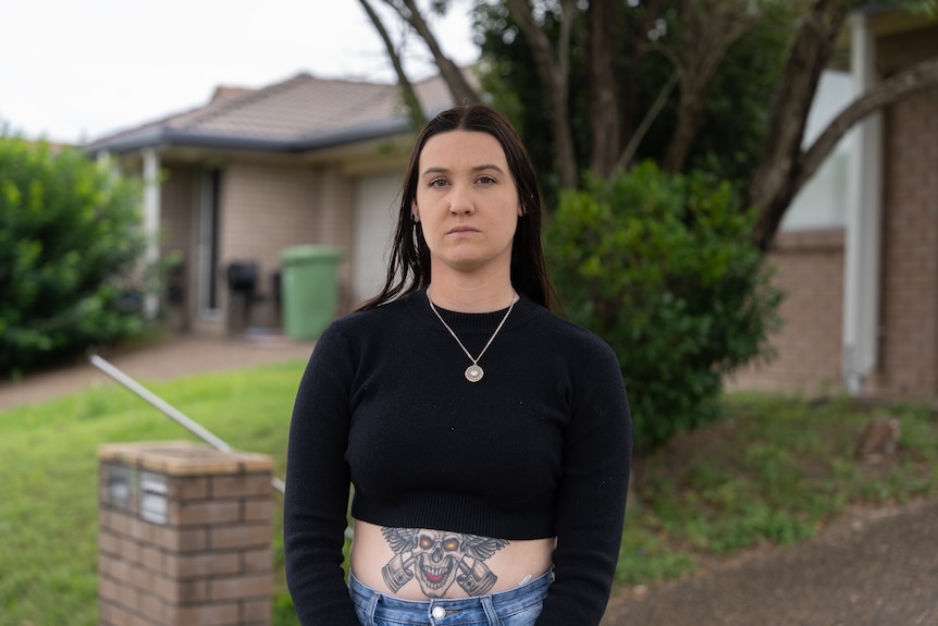 A woman stands in front of a house, looking into camera with a serious expression.