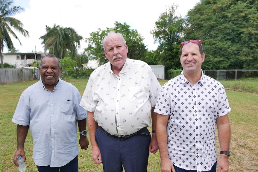 Three men in their 50s stand in an empty backyard and smile at the camera.