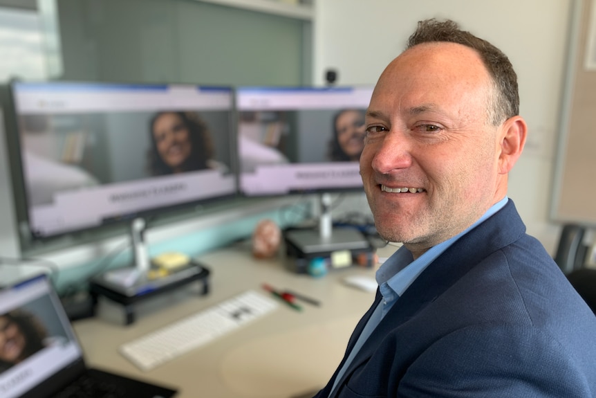 A man wearing a suit smiles as he sits at his desk, in front of a computer.