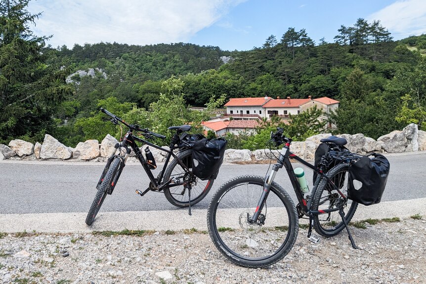 Two black bikes stand propped up on the side of the road in the countryside.