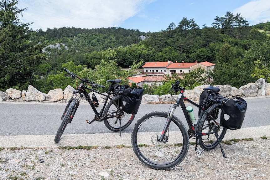 Two black bikes stand propped up on the side of the road in the countryside.