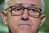 A close-up of Malcolm Turnbull's face shows him frowning. He is wearing dark rimmed glasses