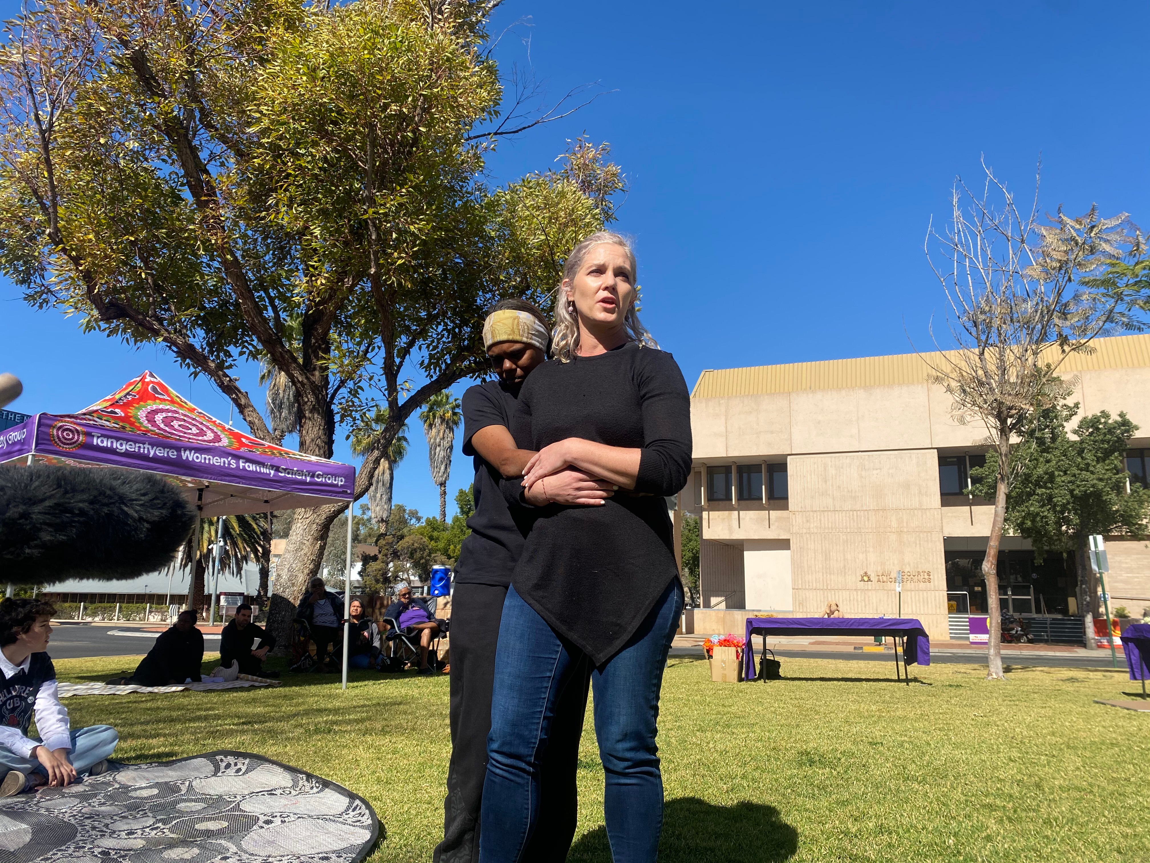 A woman stands in a grassy outdoor area speaking. Another woman is standing next to her embracing her from the side.