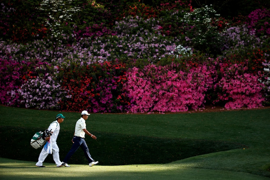 Hideki Matsuyama and his caddy walk in front of blooming pink azaleas at Augusta National