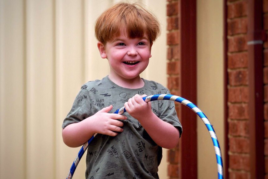 A midshot of a small boy with red hair and a green shirt standing in a back yard holding a hula hoop.