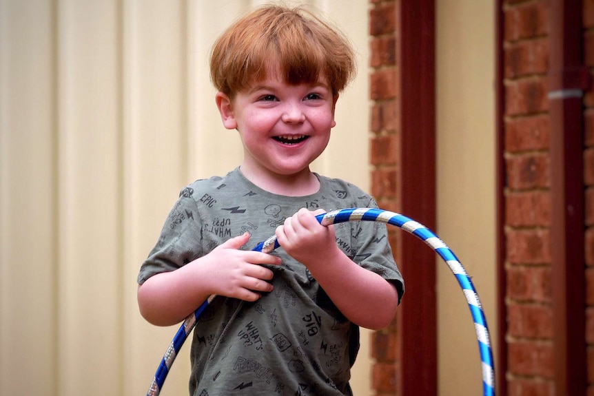 A midshot of a small boy with red hair and a green shirt standing in a back yard holding a hula hoop.