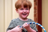 A midshot of a small boy with red hair and a green shirt standing in a back yard holding a hula hoop.