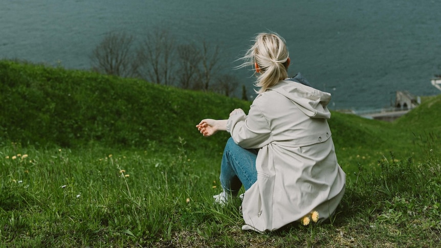 A woman with a blonde pony sits on a grassy knoll looking away from the camera to the ocean below.