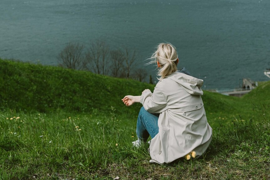 A woman with a blonde pony sits on a grassy knoll looking away from the camera to the ocean below.