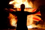 A man holds a flag in front of a raging fire on a dark night