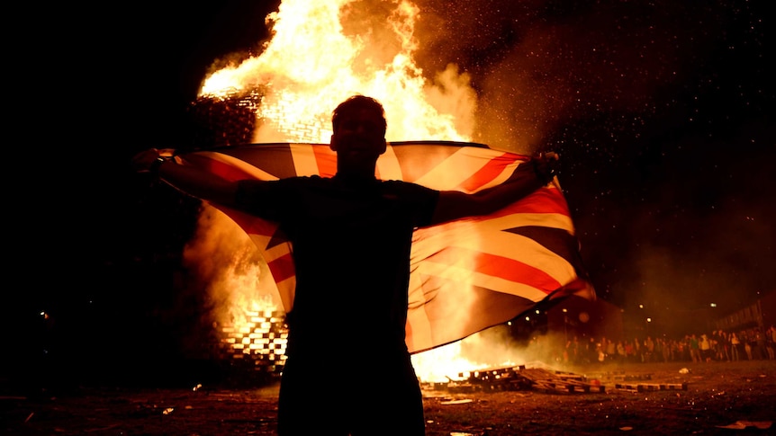 A man holds a flag in front of a raging fire on a dark night