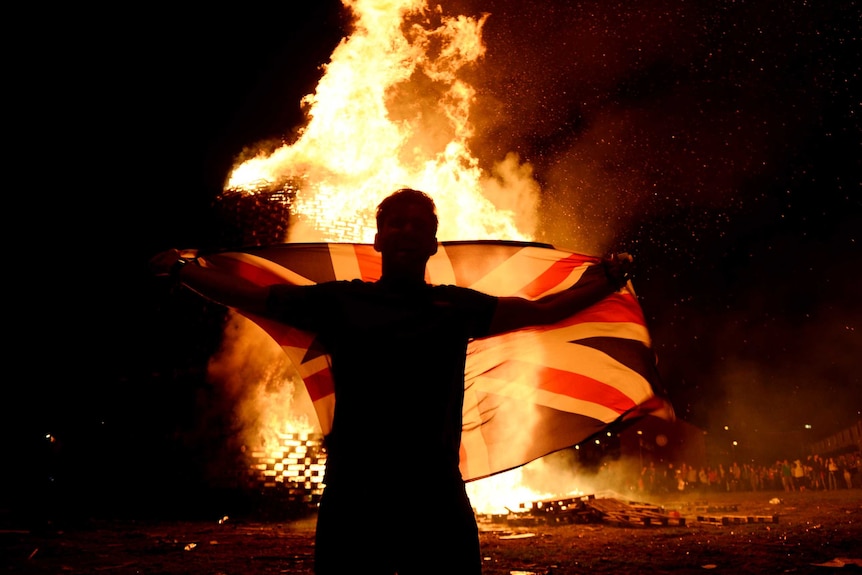 A man holds a flag in front of a raging fire on a dark night