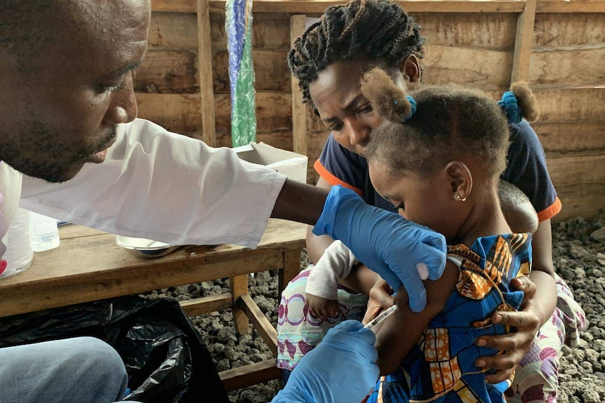 a man with gloves on inserts a needle into the arm of a young girl as a woman holds her