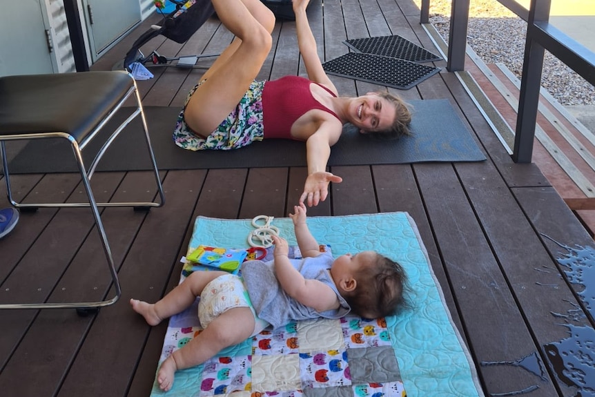 A photo of a mother lying on a yoga mat, stretching her arm out to a baby lying next to her.