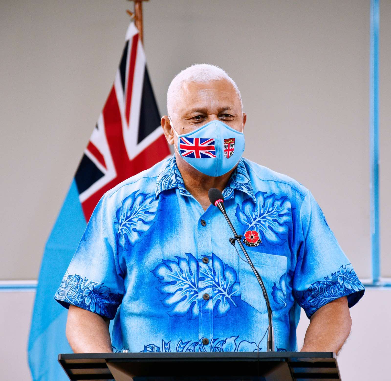 An olive skinned middle aged man with cropped white hair stands a lecturn with blue shirt and face mask in front of flag