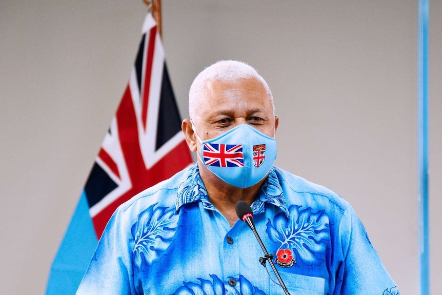 A middle-aged man in olive skin with cropped white hair stands a lecture with blue shirt and face mask in front of flag