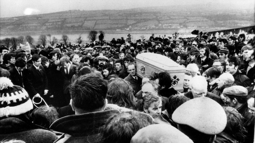 A black and white photo shows a large crowd of people and pallbearers carrying a coffin.