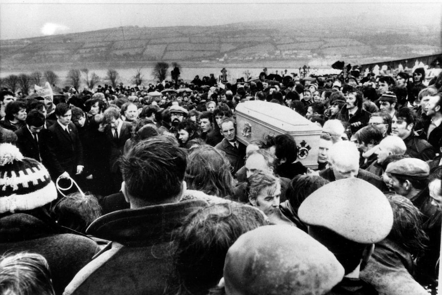 A black and white photo shows a large crowd of people and pallbearers carrying a coffin.