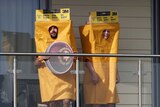 Two cricket fans wearing yellow sandpaper costumes stand on a balcony overlooking the cricket.