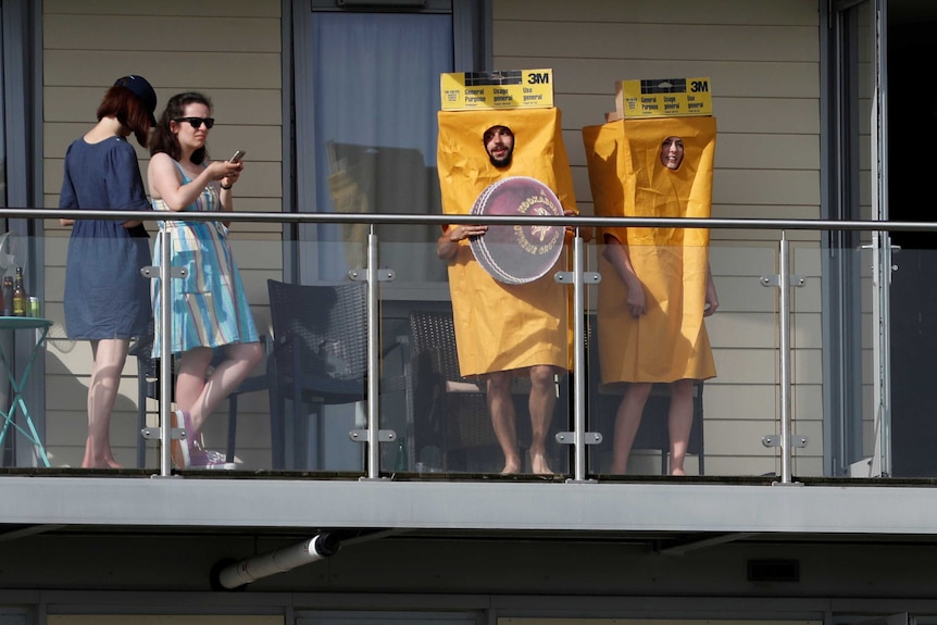 Two cricket fans wearing yellow sandpaper costumes stand on a balcony overlooking the cricket.